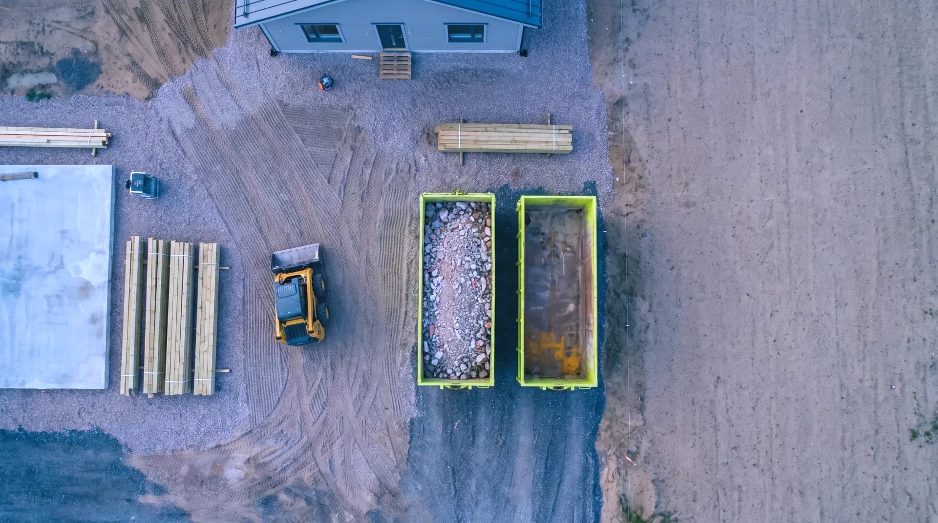 A cleared building site seen from directly above: stacked timber, a skid-steer loader and two lime containers