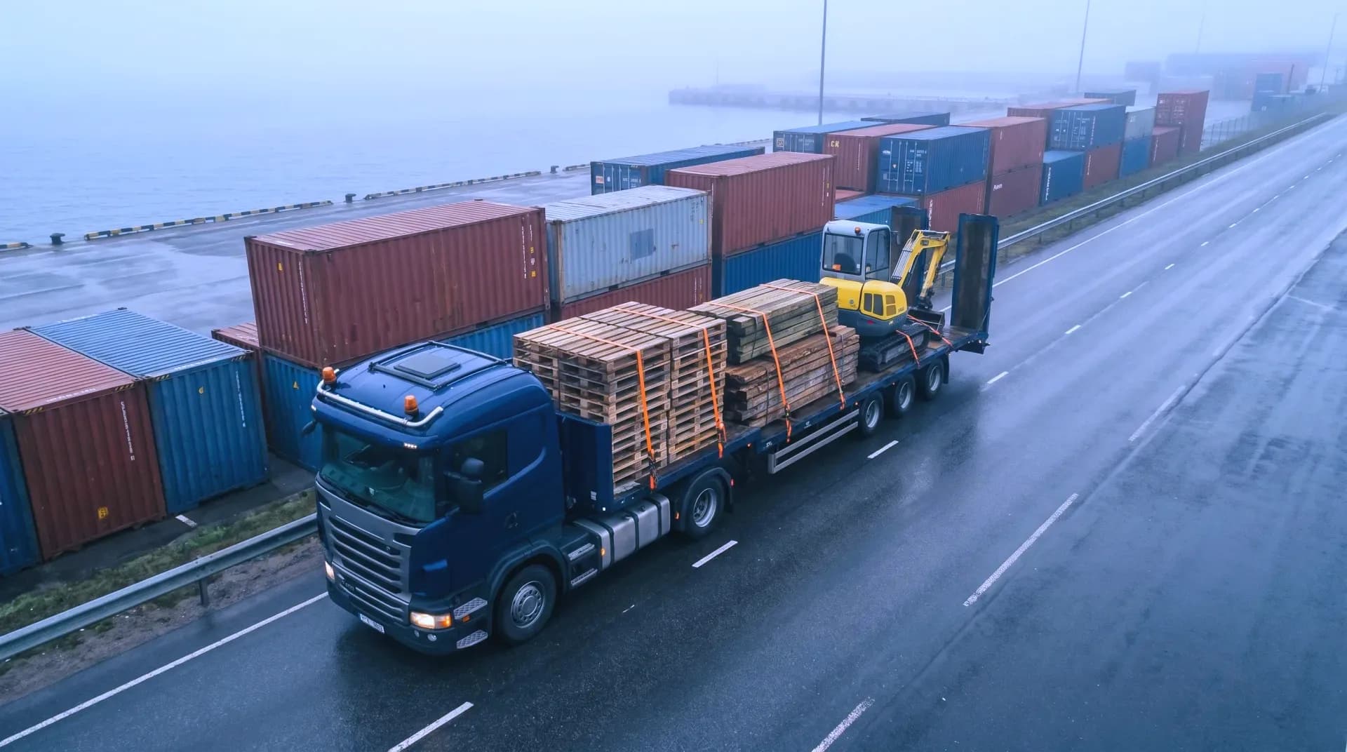 A flatbed lorry carrying strapped pallets, timber and a small excavator along a harbour road, seen from above