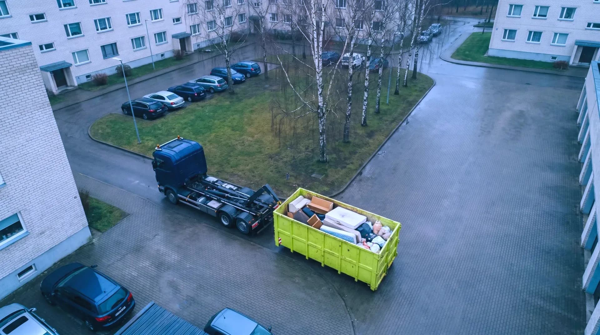 A skip lorry and a lime container filled with old furniture in the courtyard of an apartment block, seen from above